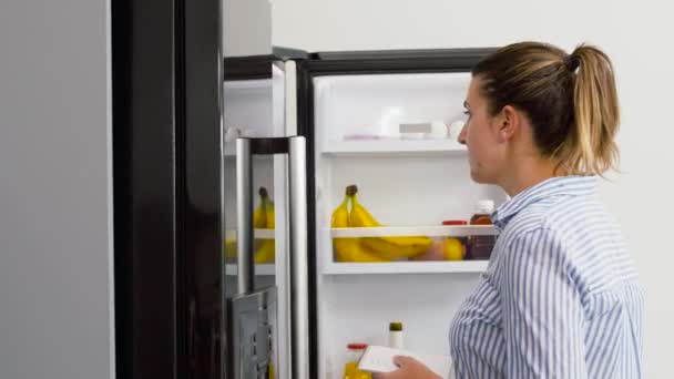 Mujer haciendo lista de alimentos necesarios en casa nevera — Vídeo de stock