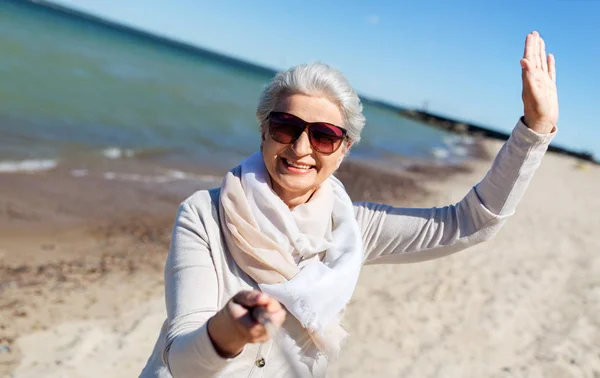 Old woman taking selfie and waving hand on beach — Stock Photo, Image