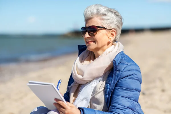 Starsza kobieta piśmie do notebooka na letniej plaży — Zdjęcie stockowe