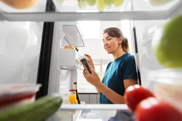 Женщина со смартфоном составляет список продуктов питания в холодильнике — стоковое фото