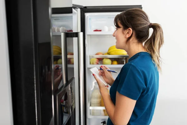 Mujer haciendo lista de alimentos necesarios en casa nevera Imagen de archivo