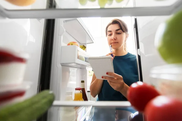 Жінка робить список необхідної їжі в домашньому холодильнику — стокове фото