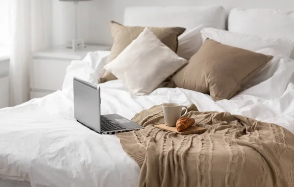 Portátil, taza de café y croissant en la cama en casa —  Fotos de Stock