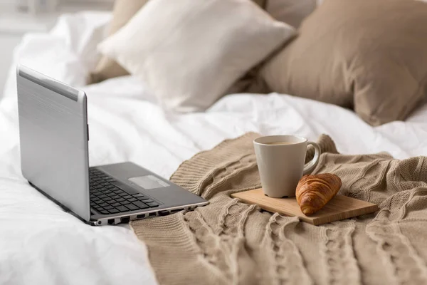 Laptop, filiżanka kawy i rogalik na łóżku w domu — Zdjęcie stockowe