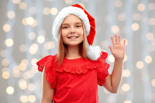 Lächelndes Mädchen mit Weihnachtsmann-Helfermütze, winkende Hand — Stockfoto