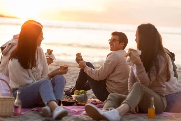 Glada vänner äter smörgåsar på picknick på stranden — Stockfoto