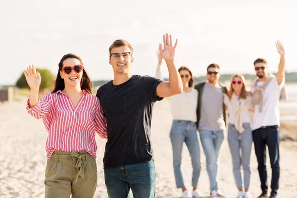 Happy couple with friends waving hands on beach — Stock Photo, Image