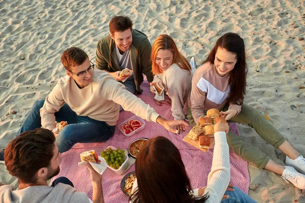 Happy friends eating sandwiches at picnic on beach — Stock Photo, Image