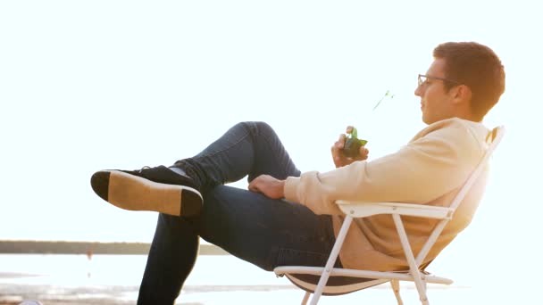 Man drinking beer sitting on chair on beach — Stock Video