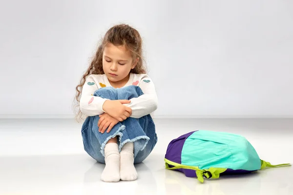 Сумна маленька дівчинка зі шкільною сумкою сидить на підлозі — стокове фото