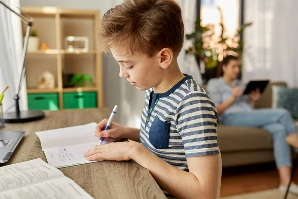 Student pojke med bok skriva till anteckningsbok hemma — Stockfoto