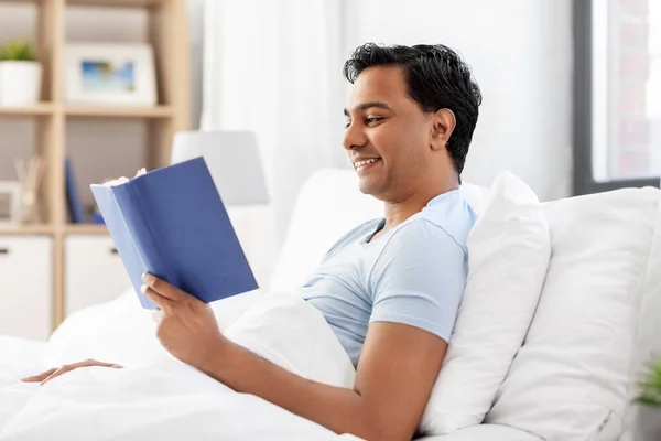 Homem indiano feliz lendo livro na cama em casa — Fotografia de Stock