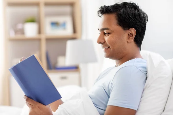 Homem indiano feliz lendo livro na cama em casa — Fotografia de Stock