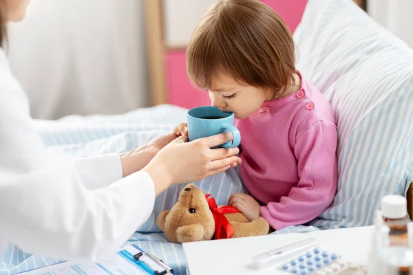 Médico dando té caliente a niña enferma en la cama —  Fotos de Stock