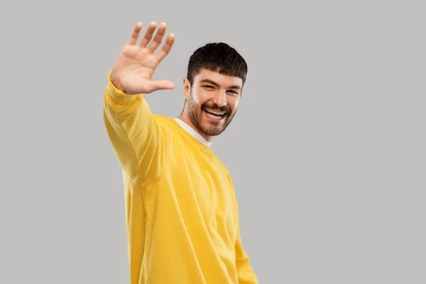 Smiling young man in yellow sweatshirt waving hand — Stock Photo, Image