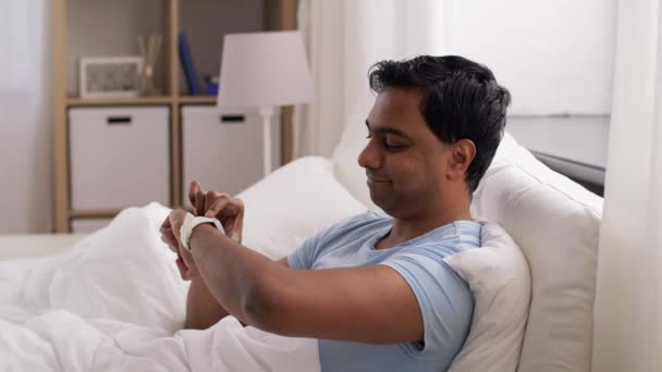 Glad indian man med smart klocka i sängen hemma — Stockvideo