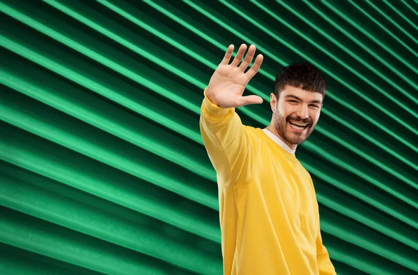 Smiling young man in yellow sweatshirt waving hand — Stock Photo, Image