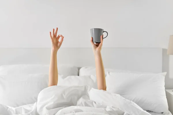 Mujer con taza de café acostado en la cama mostrando ok —  Fotos de Stock