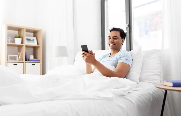Hombre indio feliz con teléfono inteligente en la cama en casa —  Fotos de Stock