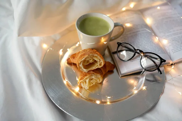 Croissant, matcha tea, könyv és szemüveg az ágyban — Stock Fotó