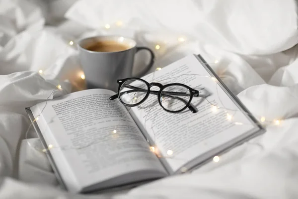 Cup of coffee, book, glasses and garland in bed — Stock Photo, Image