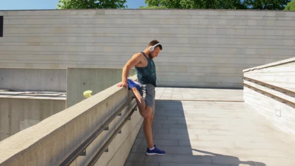 Hombre joven en auriculares que estiran la pierna al aire libre — Vídeos de Stock