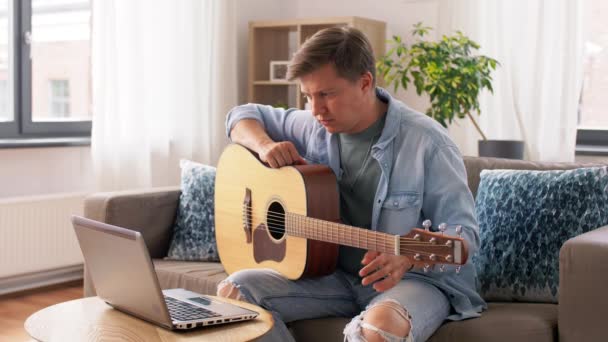 Młody człowiek z laptopem gra na gitarze w domu — Wideo stockowe