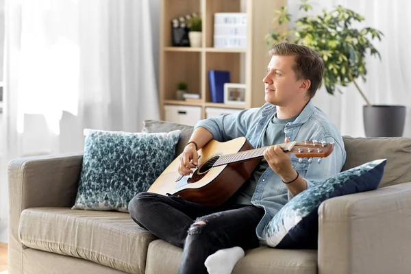 Joven tocando la guitarra sentado en el sofá en casa — Foto de Stock