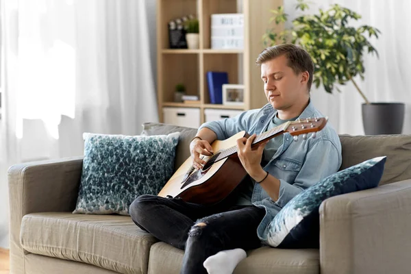 Mladý muž hraje na kytaru sedí na pohovce doma — Stock fotografie