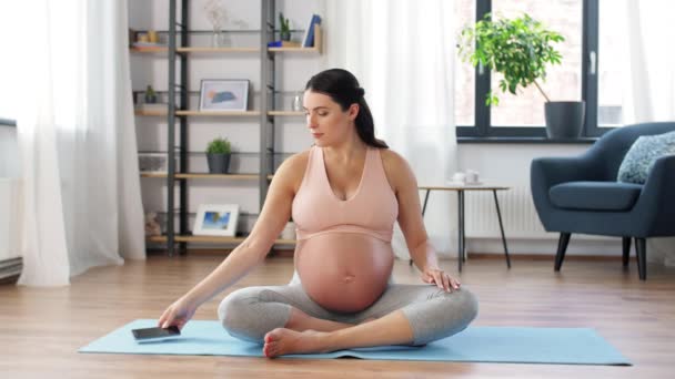 Щаслива вагітна жінка з телефоном робить йогу вдома — стокове відео