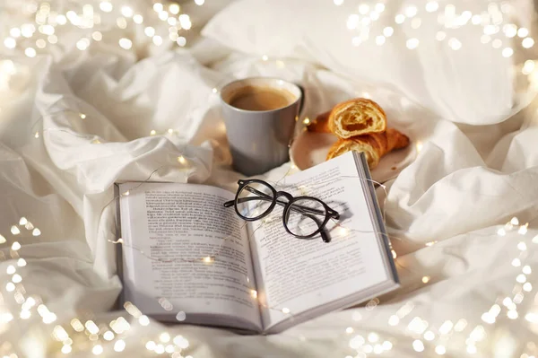 Croissant, csésze kávé, könyv és szemüveg az ágyban — Stock Fotó