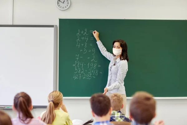 Leraar in masker schrijven op schoolbord op school — Stockfoto