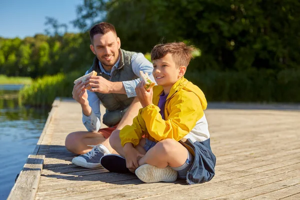 Pai e filho comendo sanduíches no cais do rio — Fotografia de Stock