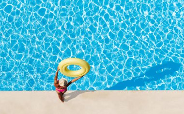 Havuz kenarında duran, ek yükü sarı şişme halka tutarak ve su yüzeyi boşluk ile seyir hasır şapka içinde kadın havadan görünümü