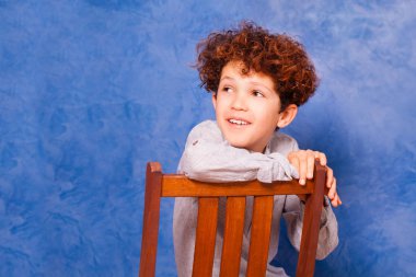 Geriye doğru boşluk ile mavi arka plan karşı ahşap sandalye üzerinde oturan varoluş kıvırcık çocuk portresi