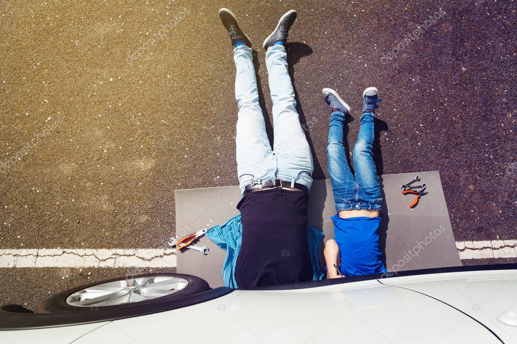 Top view picture of father and son lying down under auto underbody doing repair work on the pavement