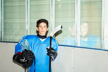 Portrait of teenage boy, professional hockey player in blue uniform, standing on ice rink, holding helmet and stick clipart