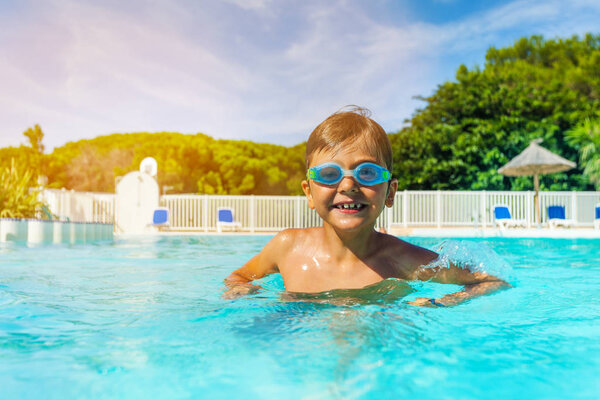 Portrait of seven years old boy wearing swimming goggles, standing in outdoor pool and looking at camera