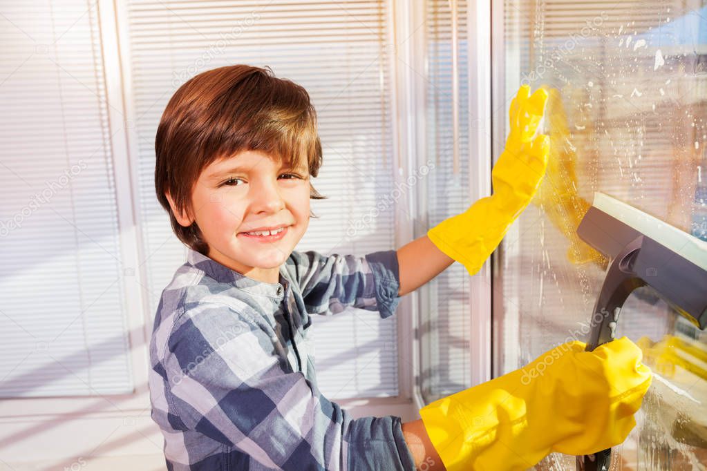 Close-up portrait of seven years old boy wearing yellow rubber gloves, washing windows with sponge and cleaning brush