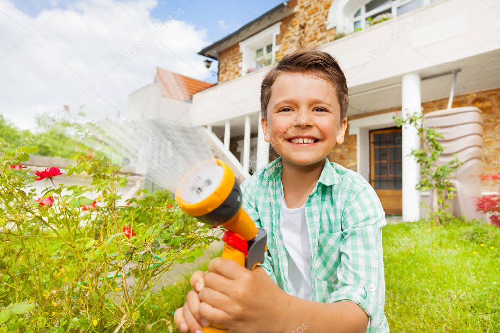 Close-up portrait of happy little boy working at garden, watering rose bushes using hand sprinkler