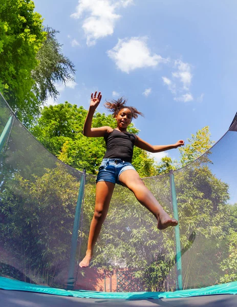 Low angle view of African girl teenager bouncing up on backyard trampoline in summer