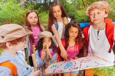 Group of kids in forest examine and point with finger on map during treasure hunting hike activity in summer camp clipart