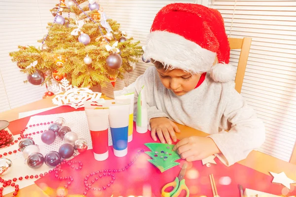 圣诞老人的帽子装饰纸圣诞树上的小男孩的肖像 坐在幼儿园的办公桌上 — 图库照片