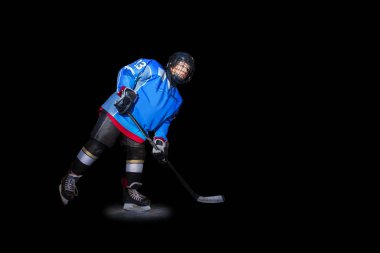 Full-length portrait of teenage boy, ice hockey player with stick in spot light over black background clipart