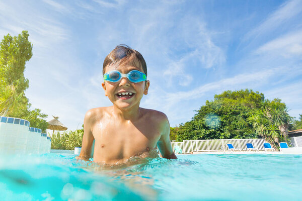 Portrait of wet boy in goggles, standing in the swimming pool, laughing and enjoying summer