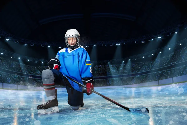 Portrait of young hockey player with stick standing on one knee on ice rink