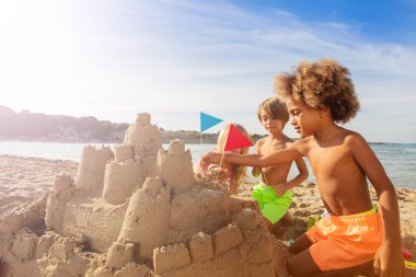 Happy friends, multiethnic boys and girls, playing at the seaside, decorating sandcastle towers with flags clipart