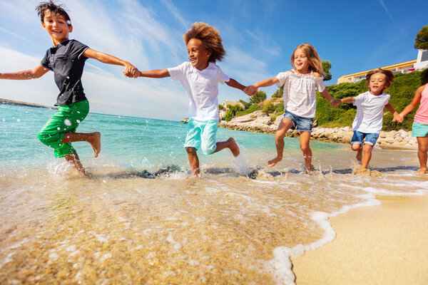 Portrait of multiethnic kids, happy friends, holding hands and running along the beach in summertime