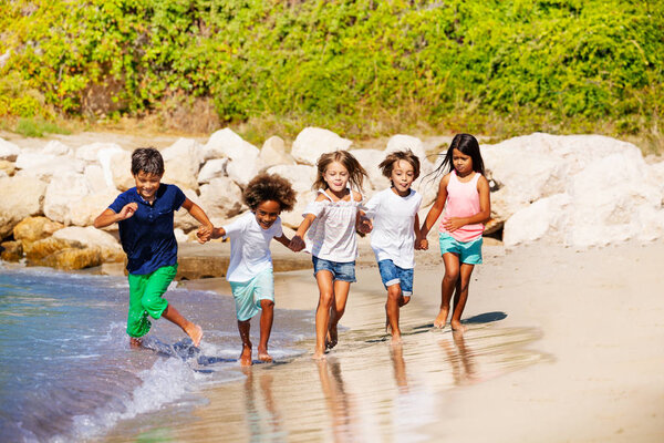 Five multiethnic kids, happy friends, holding hands and running along the beach