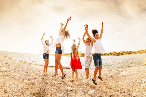 Group of excited teens, happy boys and girls, jumping in the air with hands up on the beach